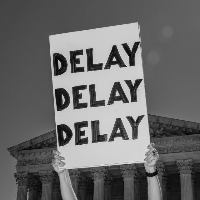 In front of the Supreme Court Building, a person holding up a sign reading, “Delay, delay, delay.”